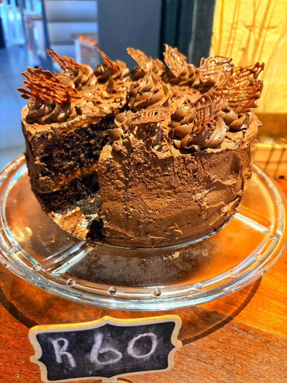 A sliced chocolate cake with piped chocolate icing and caramel, marked R60 on a small chalkboard