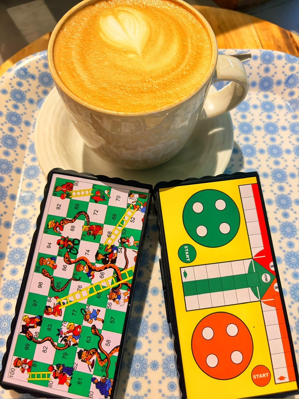 A large cappuccino with heart latte art on a tray beside snakes-and-ladders and ludo board games