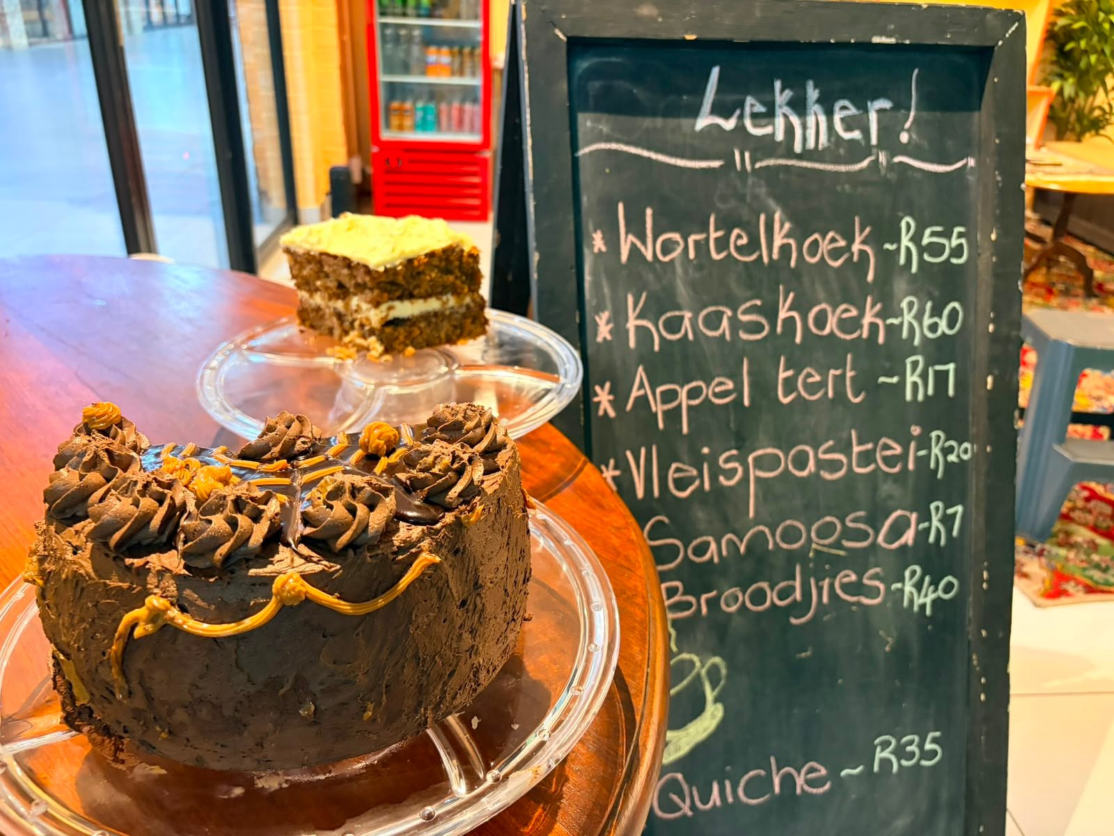 A chocolate cake and a slice of wortelkoek on glass stands on the counter at Lekker Koffiewinkel