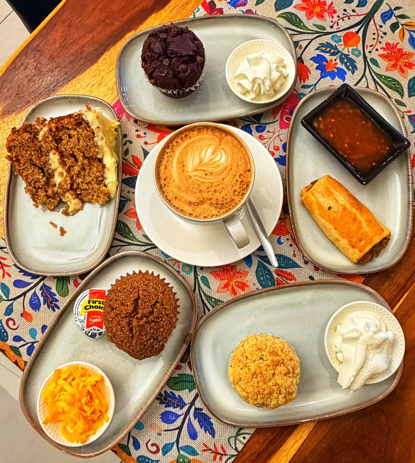 A table at Lekker Koffiewinkel laid with a cappuccino, wortelkoek, a chocolate muffin, a bran muffin with cheese, a sausage roll with chilli sauce and a melktert cup